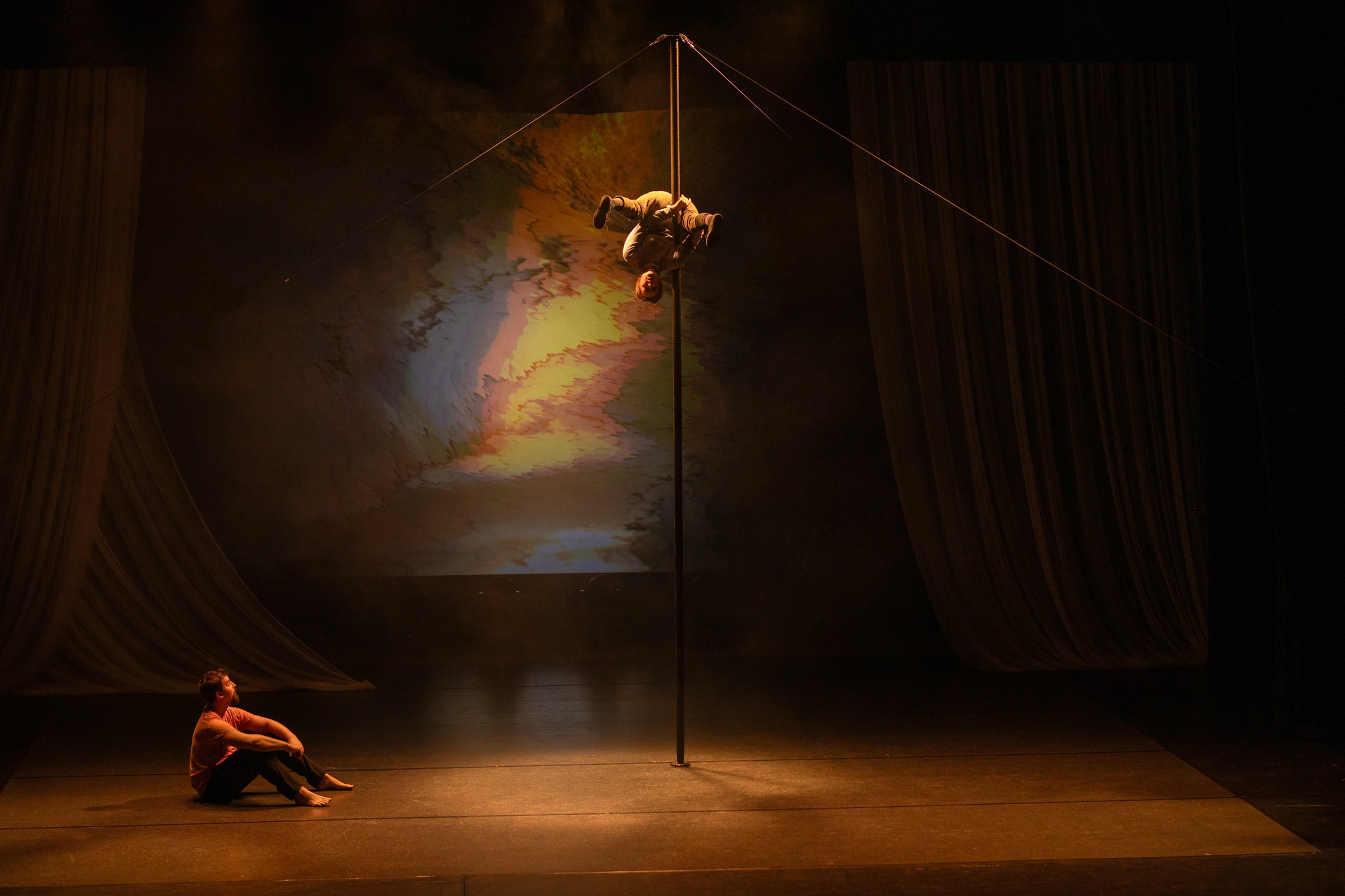 Vienas cirko artistas sėdi, o kitas atlieka triuką aukštai ant kiniško stulpo